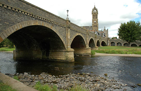 A stone bridge with multiple arches over a river, with a tower visible in the background under a cloudy sky.