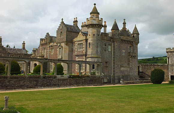 A historic castle with multiple turrets and ornate stonework, surrounded by a low stone wall, set against a backdrop of a cloudy sky and greenery.