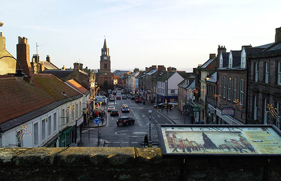 A view of a quaint town street at dusk with buildings on either side, cars on the road, and a clock tower in the distance under a clear sky.