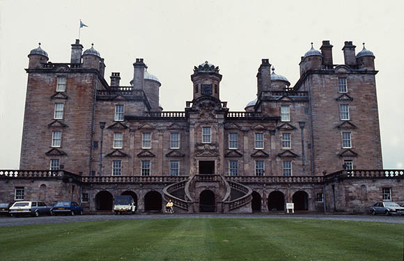 A historic stone building with turrets, a central staircase, symmetrical wings, a Scottish flag, parked cars, and a manicured lawn.