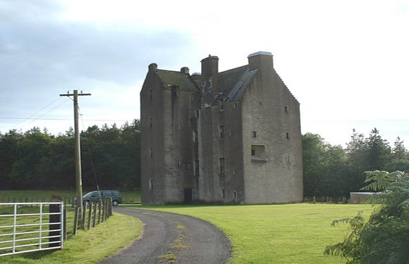 A quaint stone castle with a pitched roof is nestled in a green landscape, featuring a driveway, metal gate, and surrounding trees.