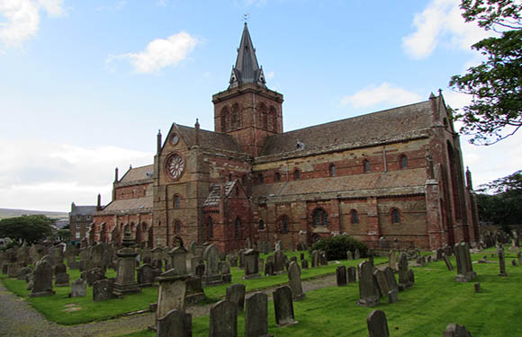 A historical stone church with a prominent clock tower and spire, surrounded by an old graveyard with numerous tombstones, under a partly cloudy sky.