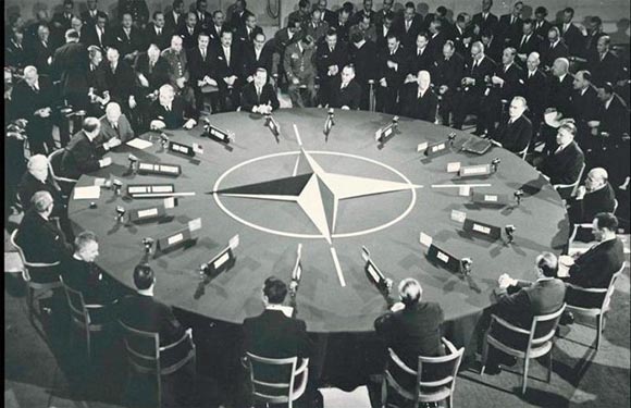 A black and white photo shows men in suits at a circular table, likely at a historical political meeting.