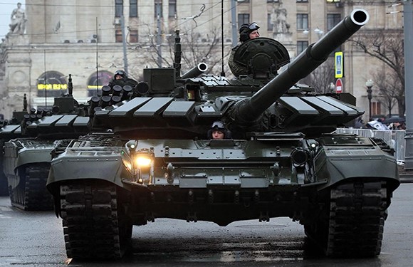 A military tank on a city street with buildings in the background, featuring a person visible in the tank's hatch.