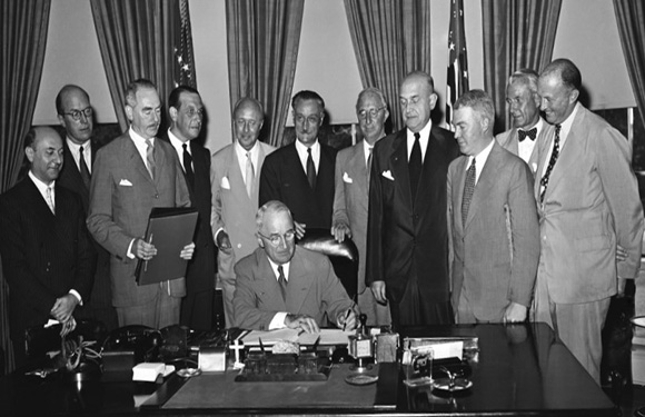 Historical photo shows a man signing a document at a desk, with a group of men and flags in a formal setting.