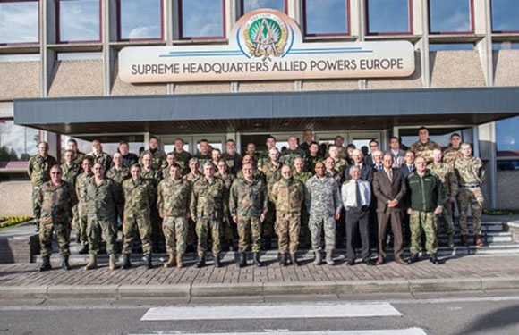 Military personnel and civilians pose for a photo in front of the "Supreme Headquarters Allied Powers Europe" building.