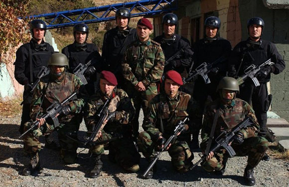 A group of military personnel in uniform, some standing and some kneeling, holding rifles, with a building and trees in the background.