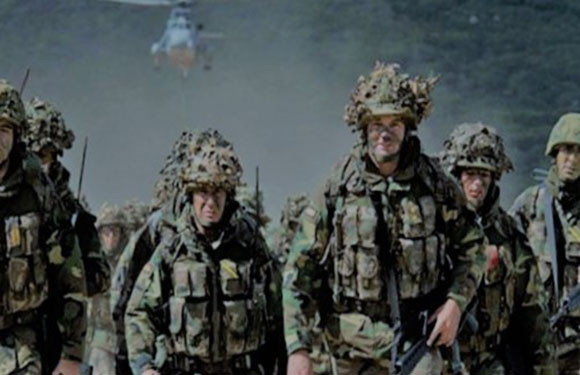 Soldiers in camouflage and helmets with foliage stand in nature, a helicopter in the background.