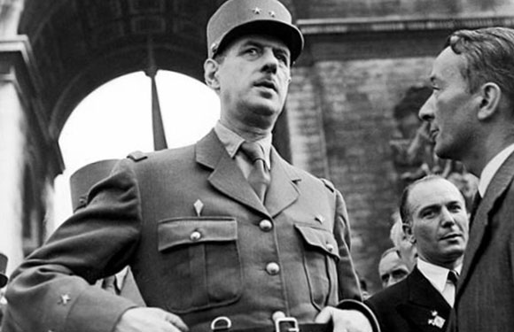 A black and white photo of a man in military uniform and cap, looking to the side with a serious expression, standing next to another man in a suit.