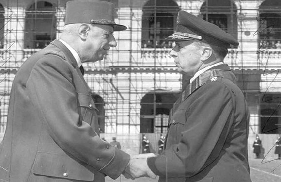 Two men in military uniforms shaking hands in front of a building with arched facades.