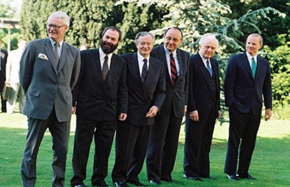 A group of six older men in suits standing outdoors, smiling for the camera with trees and a lawn in the background.