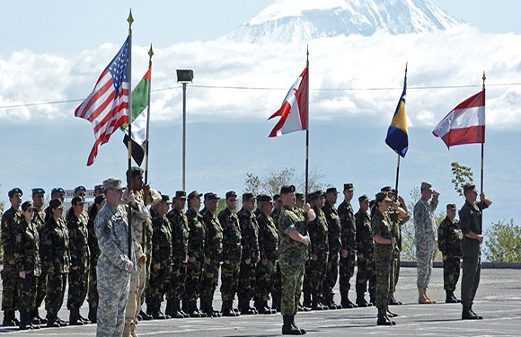 Military personnel stand in formation at a ceremony with various national flags, including the US, against a snowy mountain backdrop.