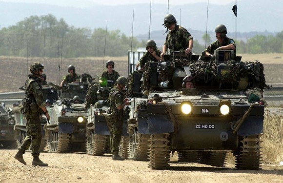 A group of military personnel in combat uniforms with helmets are positioned on and around armored vehicles in a field environment.