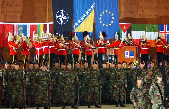 Military personnel in camouflage stand in formation as a band in red uniforms plays brass instruments, with various national and international flags displayed.