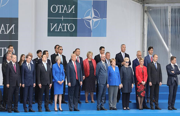 A group of formally dressed individuals standing in a row for a photo op in front of a backdrop with the NATO logo and Cyrillic text.