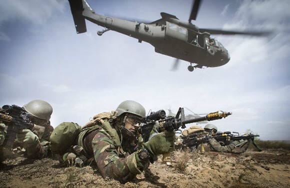 A group of soldiers in combat gear lying prone on the ground with rifles aimed, with a helicopter flying low in the background.