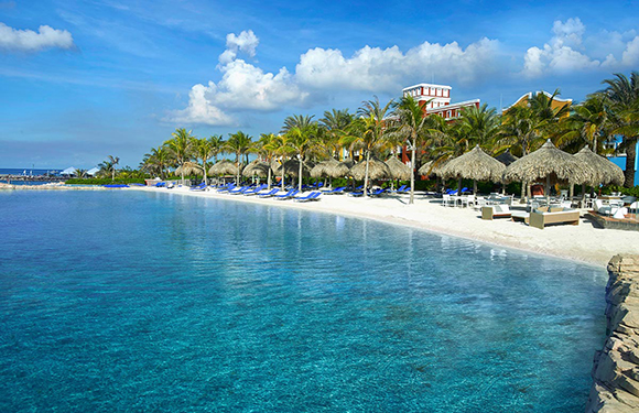 A tropical beach resort with clear blue water, palm trees, sun loungers, and thatched umbrellas along the shoreline, under a partly cloudy sky.