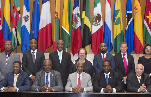 Formally dressed people posing for a photo in front of international flags, with one row seated and one standing.