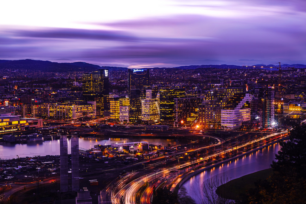 A cityscape during twilight with illuminated buildings and a river reflecting city lights, under a sky with streaks of purple and blue clouds.