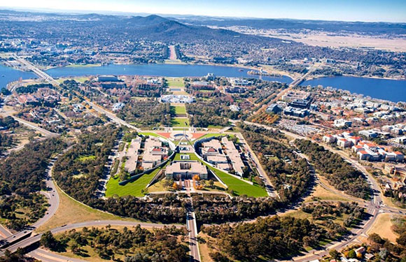 Aerial view of a planned city with symmetrical layout, featuring roads, buildings, and green spaces, with a lake in the background.