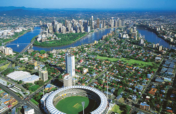 Aerial view of a city with skyscrapers near a winding river, a stadium in the foreground, and mountains in the distance.