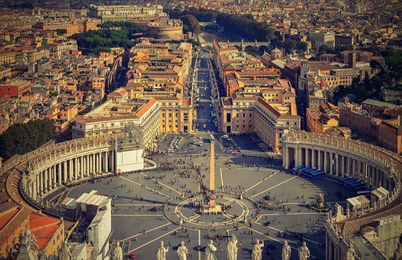 Aerial view of St. Peter's Square in Vatican City, showcasing the obelisk, colonnades, and surrounding buildings with a cityscape in the background.