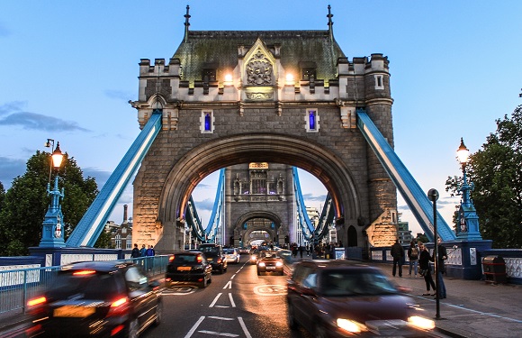 A photograph of Tower Bridge in London during twilight with vehicle traffic and the bridge's lights illuminated.