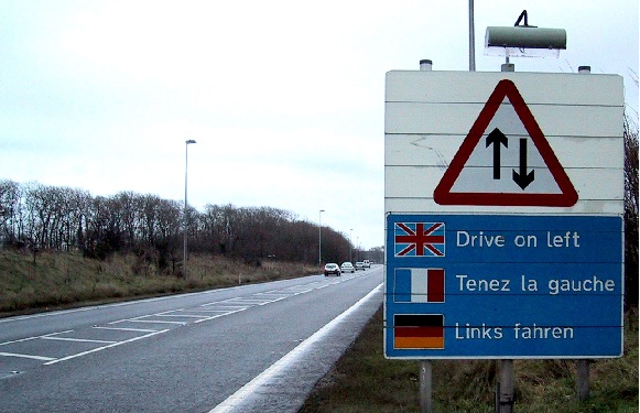 A multilingual traffic sign indicates two-way traffic and instructs to drive on the left, with an empty road behind.