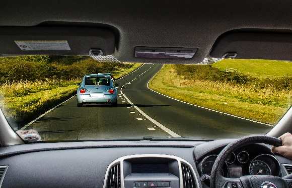 A view from the inside of a car showing another car ahead on a curving road surrounded by greenery.