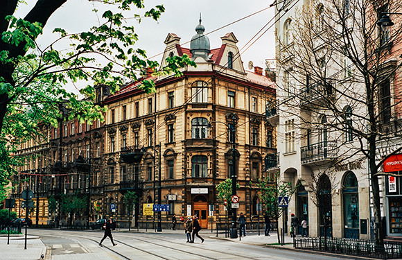 A street view with people walking, featuring a corner building with ornate architecture, a tram line, and green trees.