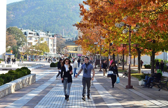 A couple strolls on a leafy walkway, with others on benches and a mountain backdrop.