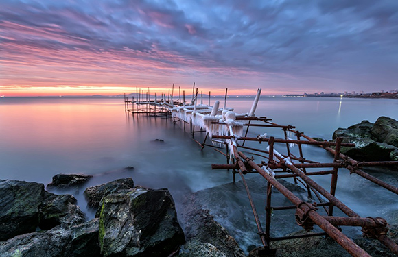 A peaceful twilight over a calm lake with a sky shifting from pink to blue, featuring a broken pier and visible rocks.