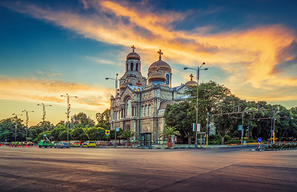 An image of a grand cathedral with multiple domes under a dramatic sky at sunset, with surrounding greenery and a street in the foreground.