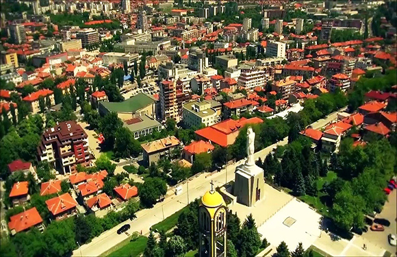 Aerial view of a cityscape with dense building clusters, green trees, and a prominent monument in the foreground.