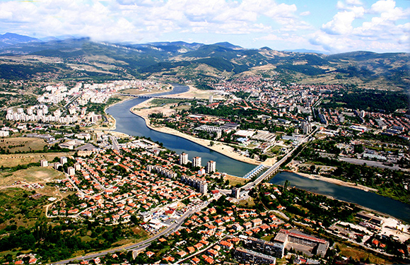 Aerial view of a city with a river curving through it, bridges spanning the river, and various buildings and green areas visible.