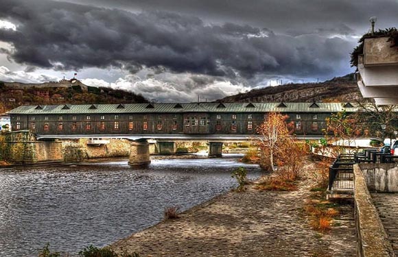 A covered bridge with multiple arches spans across a river under a cloudy sky. Trees and a building are visible on the riverbank.