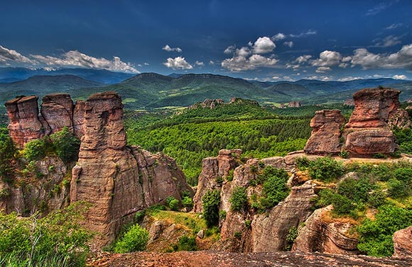 A picturesque landscape with tall rock formations, greenery, a lush valley, distant mountains, and a partly cloudy sky.