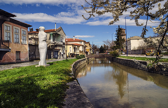 A scenic canal view with clear water, European buildings, lush greenery, a statue, and blossoming trees.