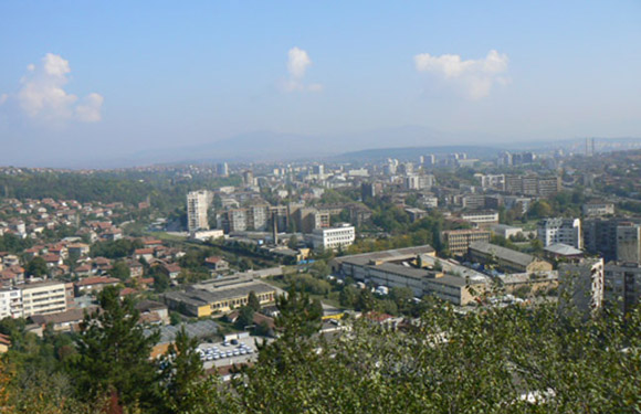 A panoramic view of a cityscape with various buildings, surrounded by greenery and trees, under a partly cloudy sky.