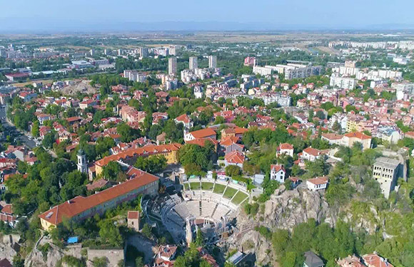 Aerial view of a city with buildings, streets, and greenery, featuring an amphitheater in the foreground.