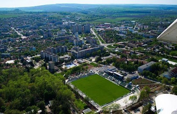 Aerial view of a suburban area with residential buildings, a soccer field, and greenery, with a cityscape and hills in the distance.