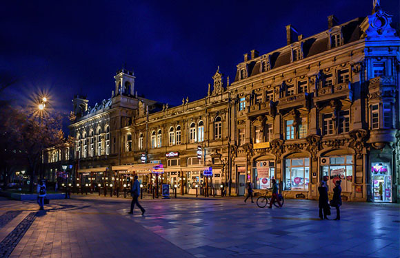 A nighttime cityscape with illuminated buildings and a few pedestrians on a wide sidewalk.