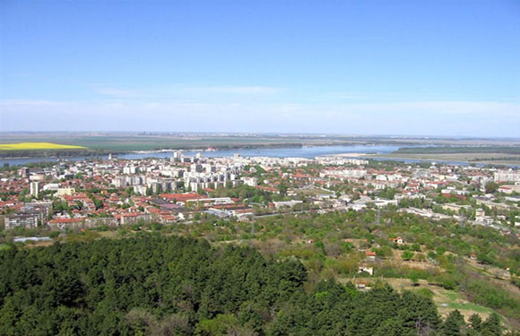 Aerial view of a town with buildings and green areas, with a body of water in the distance and a clear sky above.