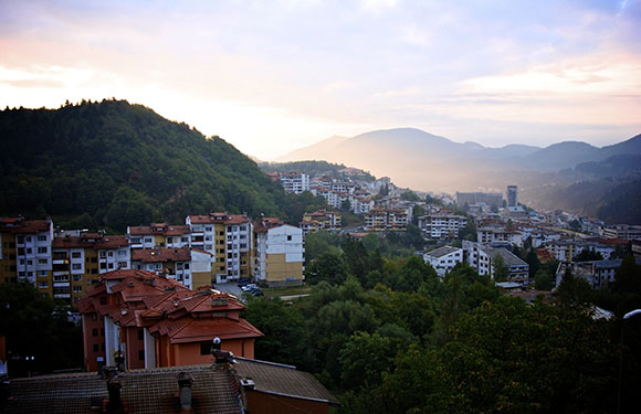 A scenic view of a town nestled in a valley with buildings scattered across the hillsides, under a sky with soft light, possibly at dawn or dusk.