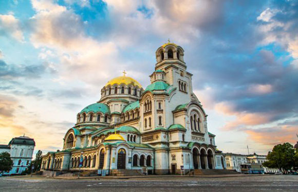 An image of the Alexander Nevsky Cathedral with a cloudy sky at sunset or sunrise.