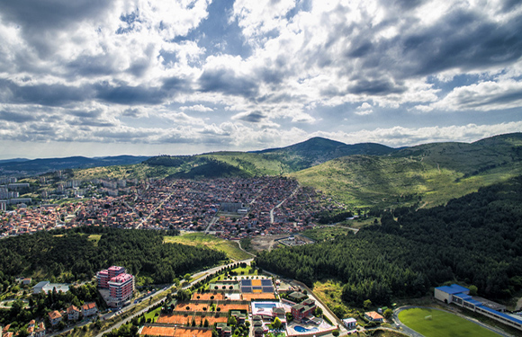 Aerial view of a town nestled in a valley with surrounding green hills and scattered clouds above.