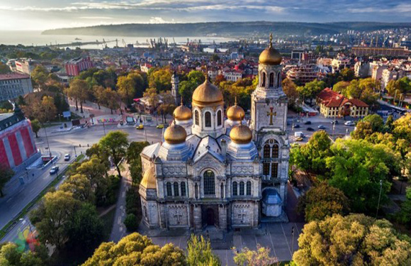 Aerial view of a large ornate church with golden domes, surrounded by trees, with a cityscape and water in the background under a partly cloudy sky.