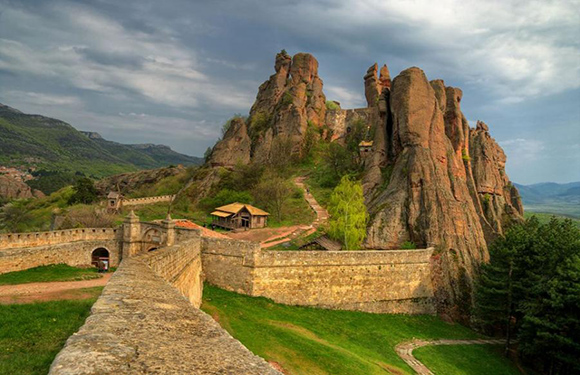 A scenic view of a historic fortress with tall, rocky cliffs and a stone wall leading up to a gate, surrounded by lush greenery under a cloudy sky.