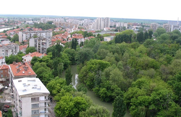 Aerial view of a cityscape with buildings surrounded by green areas and trees, with a river running through the foliage.