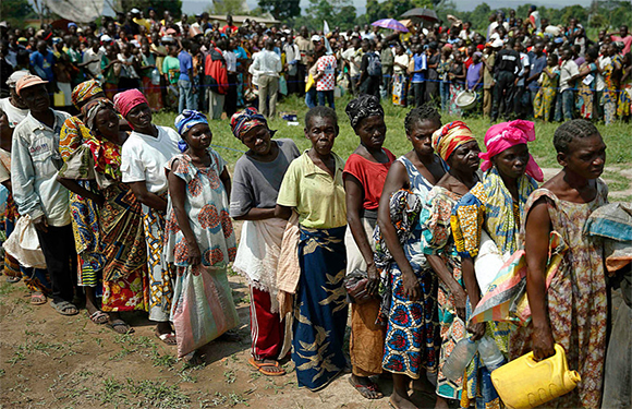 A group of people standing in a line outdoors, wearing various colorful clothes, with more individuals and trees visible in the background.